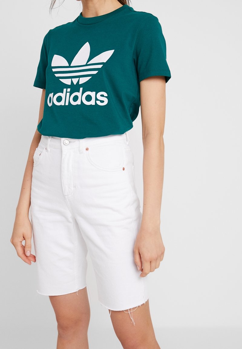 Camiseta de algodón en color verde azulado con logo blanco de Adidas y diseño de tres rayas, combinada con pantalones cortos de mezclilla blanca de cintura alta con bordes deshilachados.