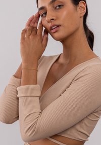 Beige gestreiftes Langarmoberteil mit tiefem V-Ausschnitt und umgeschlagenen Manschetten. Ein dünnes goldenes Armband schmückt das Handgelenk und fügt eine Detail hinzu.