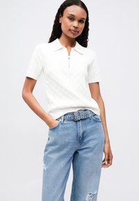 Fehér, texturált, rövid ujjú pólóing cipzáros gallérral, világoskék, megviselt, magas derekú farmernadrággal és fekete övvel párosítva.