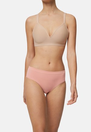 Mujer con un sujetador beige sin costuras y ropa interior rosa de corte alto, contra un fondo claro y liso.