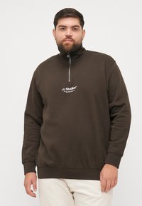 Sweatshirt zippé marron avec col montant, arborant un logo blanc sur la poitrine. Manches longues et texture douce, terminé par un ourlet côtelé.