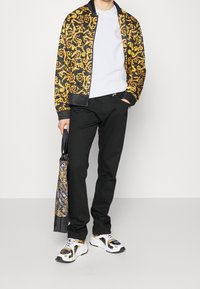 Giacca bomber nera con motivo a spirale dorata, t-shirt bianca, pantaloni neri e sneakers nere e dorate. Tiene una borsa a tracolla abbinata.