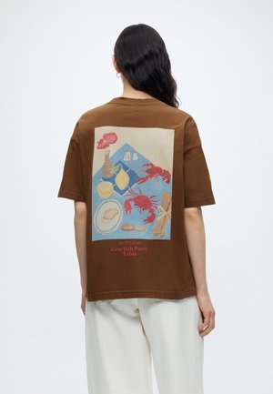 Άτομο με μακριά σκούρα μαλλιά φοράει καφέ μπλουζάκι με πολύχρωμη εικονογράφηση δείπνου με καρκινοειδή και λευκό παντελόνι, με την πλάτη γυρισμένη.