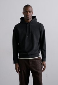 Svart hoodie med texturerad tyg, dragsnören och en tapered nederkant, bärs över mörka randiga byxor med en kontrasterande ljusfärgad midja.