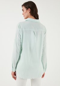 Light mint green, long-sleeved shirt with a collar, back pleat, and frayed hem. Made from textured fabric, features buttoned cuff details.