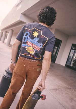 Mand med krøllet hår, der bærer skateboard, går på fortovet nær bygningskolonner iført en mærket t-shirt og brune bukser.