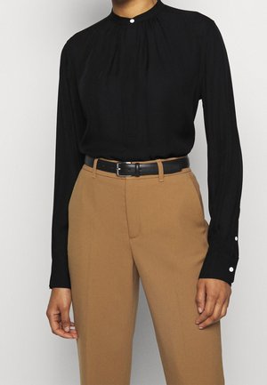 Osoba nosząca czarną bluzkę z długim rękawem z guzikami i wysokim stanem camelowe spodnie zabezpieczone czarnym skórzanym paskiem.