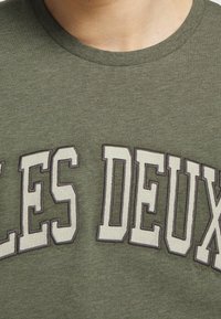 Zielona koszulka z krótkim rękawem wykonana z miękkiego materiału, z białym haftowanym napisem "LES DEUX" w grubym kroju na klatce piersiowej.