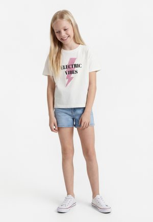 Mosolygós lány hosszú szőke hajjal, fehér "ELECTRIC VIBES" feliratú pólóban, világoskék farmer rövidnadrágban és fehér tornacipőben, fehér háttér előtt áll.