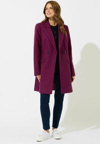 Cappotto doppiopetto in ricco misto lana viola, con revers a scampanatura, tasche frontali e silhouette dritta, abbinato a jeans scuri e sneakers bianche.