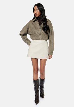 Kvinde iført en olivengrøn cropped jakke over en sort rullekrave, en hvid mininederdel og sorte knæhøje spidse støvler, stående med en hånd på hoften.