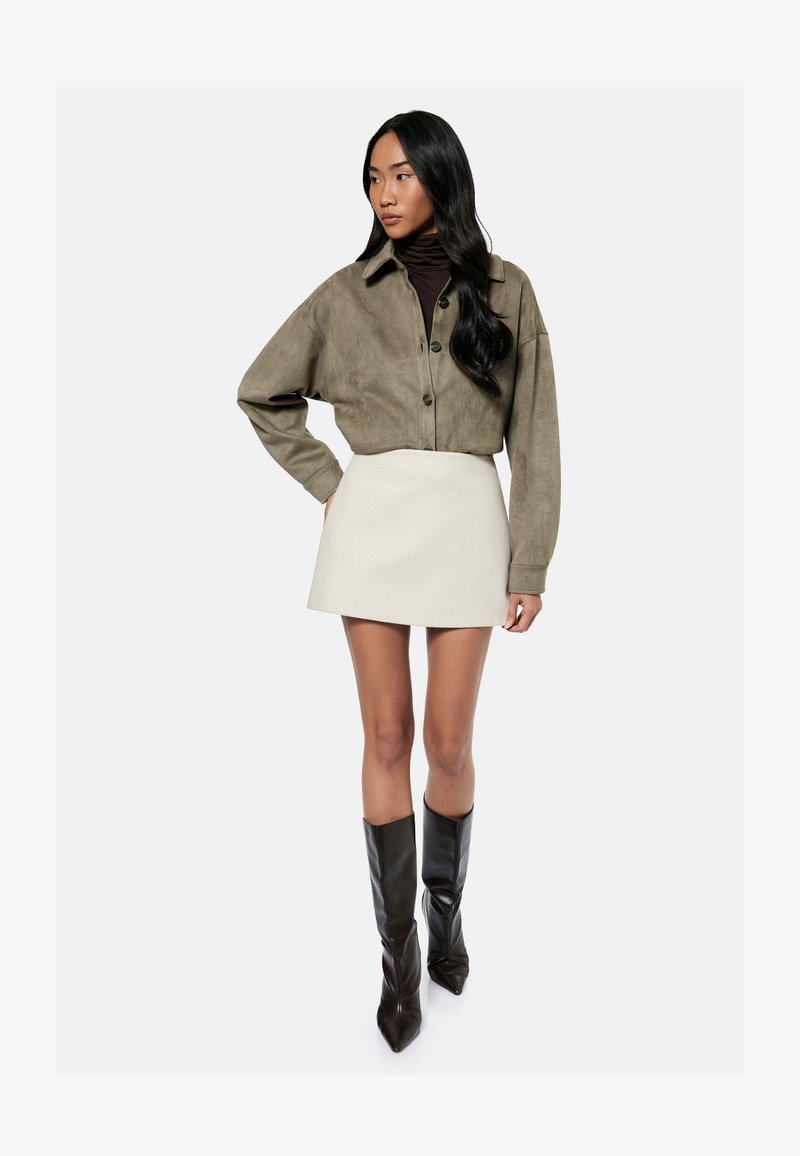 Frau trägt eine olivgrüne kurze Jacke über einem schwarzen Rollkragenpullover, einen weißen Minirock und schwarze kniehohe spitze Stiefel, steht mit einer Hand in der Hüfte.