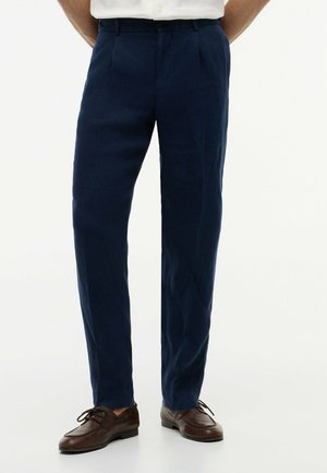 Mężczyzna w granatowych, dopasowanych spodniach i brązowych skórzanych eleganckich butach na sznurowadła, stoi z rękami za plecami.