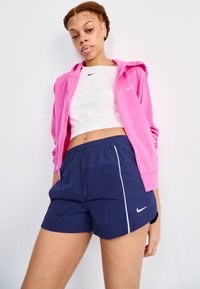 Rožinė užtrauktuku užsegama striukė virš balto trumpaoodžio, derinama su tamsiai mėlynomis šortais su balta šonine juostele ir elastiniu juosmeniu. Matomi Nike logotipai.