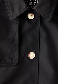 Czarna, lekka koszula z materiału z wywiniętym kołnierzykiem, z dużym, kremowym guzikiem i kieszenią po lewej stronie.