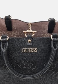 Sac à main texturé noir en matériau synthétique, avec des ferrures dorées, un intérieur à motifs, et le logo "GUESS" à l'avant.