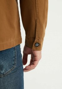 Bras droit dans une chemise à manches longues marron avec un poignet boutonné à côté de la poche d'un jean en denim bleu sur un fond clair.