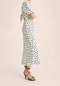 Robe blanche à pois avec une silhouette ajustée, des manches bouffantes, un dos nu et un détail à nouer. Associée à des talons noirs à lanières.