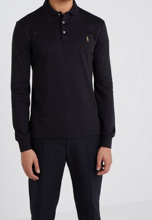 Mand iført sort langærmet poloshirt med lille broderet logo på brystet og sorte teksturerede bukser, stående mod hvid baggrund.