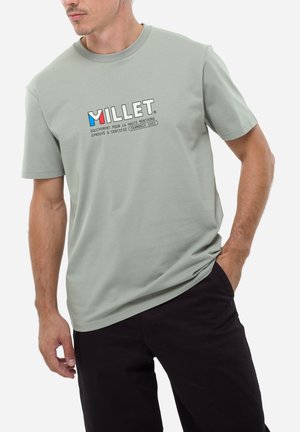 Homme portant un t-shirt Millet gris clair avec un logo bleu, blanc et rouge, et un pantalon noir, debout avec une main dans la poche.