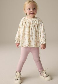 Langärmliges Oberteil mit weißem Blumenmuster und gerafftem Ausschnitt, kombiniert mit hellrosa Skinny-Hosen und glitzernden High-Top-Sneakern.