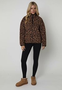 Brązowa bluza polarowa w panterkę z czarnym zamkiem błyskawicznym, luźny krój, zestawiona z czarnymi legginsami i beżowymi botkami za kostkę. Miękka faktura.