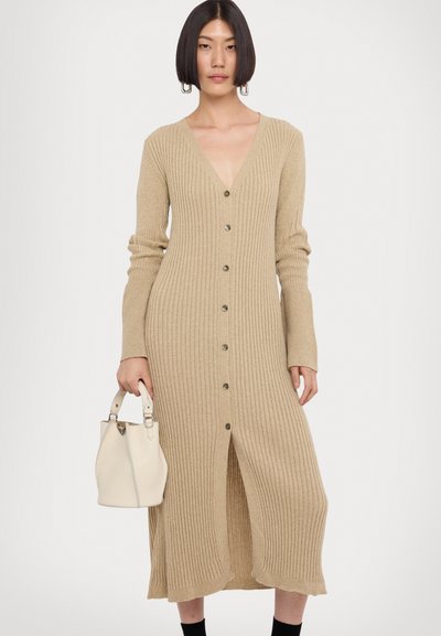 Beige ribbelgebreide jurk met een V-halslijn, knopen aan de voorkant en lange mouwen. Opvallende split aan de zoom, gecombineerd met een crèmekleurige emmerzak.