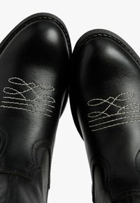 Des chaussures en cuir noir avec une broderie blanche complexe, un bout pointu et des coutures décoratives le long des bords.