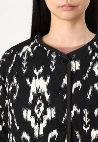 Casaco acolchoado em preto e branco com um padrão abstrato, gola redonda e três botões pretos na frente.