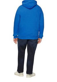 Niebieska bawełniana bluza z kapturem ze sznurkiem, noszona z ciemnymi dżinsami i jasnymi trampkami, widok od tyłu.