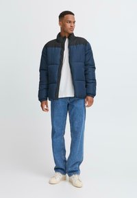 Polstret marineblå jakke med sorte skuldre, lynlås-lukning og elastiske manchetter, kombineret med lyseblå jeans og cremefarvede sneakers. Glat tekstur.