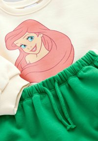 Sweat-shirt crème avec un graphique rose d'un personnage ; pantalon de survêtement vert avec une taille élastique et un cordon de serrage, texture de tissu douce.