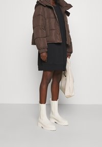 Braune, kurz geschnittene Pufferjacke mit Kapuze, oversized schwarzes Kleid, weiße klobige Stiefel und eine hellbeige Tasche. Glatte Texturen durchgehend.