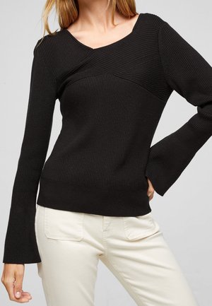 Pull à manches longues noir en jersey avec un design unique en point de croix au niveau du col et des manches évasées, associé à un pantalon couleur crème.