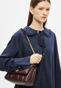 Μπλούζα ναυτικού μπλε με γιακά με βολάν, μακριά μανίκια και φιόγκο. Κρατώντας μια μπορντό δερμάτινη τσάντα χειρός με χρυσή αλυσίδα.