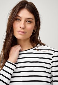 Witte top met lange mouwen en zwarte horizontale strepen; zachte stof; ronde halslijn; model houdt haar hand bij de hals.