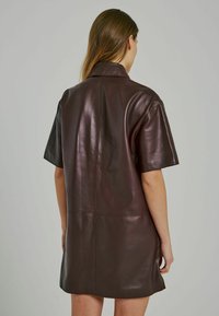 Abito in pelle marrone con maniche corte, colletto a punta, texture liscia e silhouette dritta, con una cucitura centrale e un design minimalista.