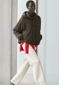 Frau geht drinnen und trägt eine braune Kapuzenjacke, weiße weite Hosen und einen roten Pullover, der um ihre Taille gebunden ist.