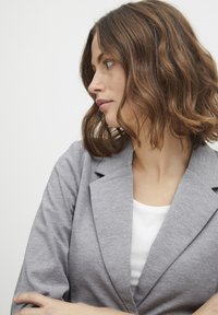 Grå blazer med texturerad yta, med kavajkrage och avslappnad passform, bärs över en vit topp. Frisyr är vågig och axellång.