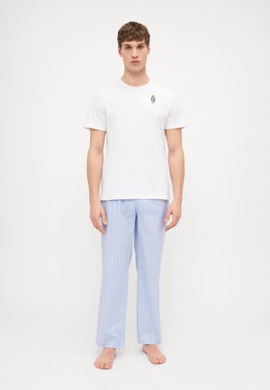 Hvid bomulds t-shirt med lille blå grafik. Lyseblå stribede pyjamabukser, lige pasform. Modellen står barfodet på en neutral baggrund.