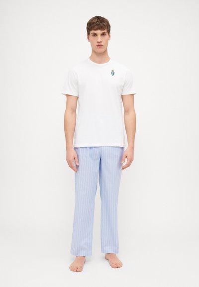 T-shirt en coton blanc avec un petit graphisme bleu. Pantalons de pyjama rayés bleu clair, coupe droite. Le modèle est pieds nus, debout sur un fond neutre.