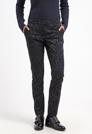 Persoon draagt strakke zwarte broek met subtiele luipaardprint, gecombineerd met een donker geribd truitje en zwarte glanzende schoenen, handen in de zakken.