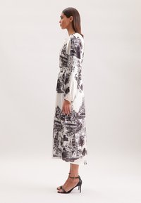 Langes weißes Kleid mit schwarzen künstlerischen architektonischen Drucken, langen Ärmeln, einer taillierten Taille und einer fließenden Silhouette. Kombiniert mit schwarzen, schmalen Absatzsandalen.