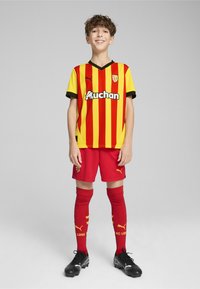 Maglia da football a righe rosse e gialle con accenti neri, pantaloni corti rossi e calze rosse con dettagli gialli. Le scarpette nere completano l'outfit.