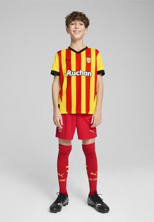 Maglia da football a righe rosse e gialle con accenti neri, pantaloni corti rossi e calze rosse con dettagli gialli. Le scarpette nere completano l'outfit.