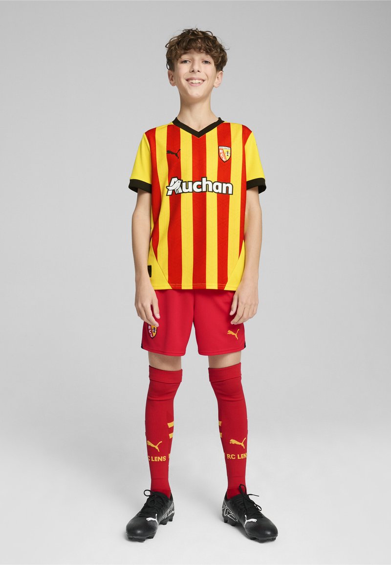 Maglia da football a righe rosse e gialle con accenti neri, pantaloni corti rossi e calze rosse con dettagli gialli. Le scarpette nere completano l'outfit.