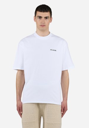 Hombre que lleva una camiseta blanca lisa de manga corta con un pequeño logo negro "PEGADOR" en el pecho izquierdo, combinado con pantalones beige texturizados.