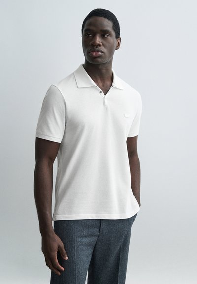 Homme portant un polo à manches courtes blanc et un pantalon gris, debout avec les mains partiellement dans les poches, regardant légèrement vers la droite.