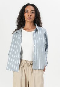  Lichtblauw overhemd met witte verticale strepen, voorzien van een borstzak, gedragen over een witte geribbelde tanktop en beige broek.
