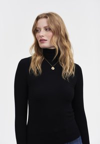 Femme aux cheveux roux ondulés portant un col roulé noir et un pendentif en or, regardant de côté sur un fond clair uni.